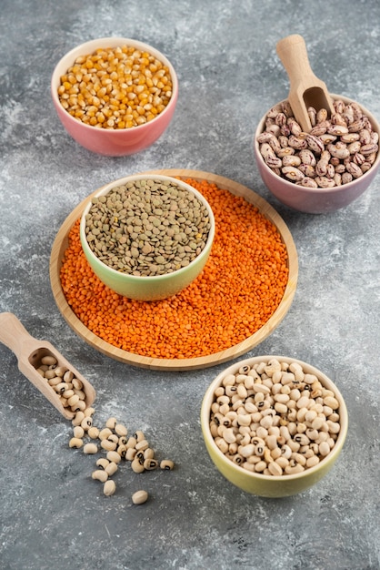 Colorful bowls of various uncooked beans, lentils and corns on marble table surface.