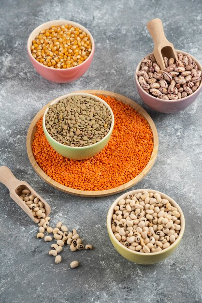 Colorful bowls of various uncooked beans, lentils and corns on marble table surface.