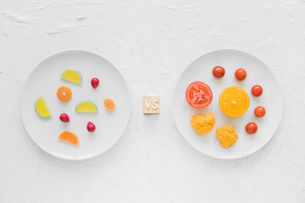 Colorful candies versus fresh vegetables on white plate over background