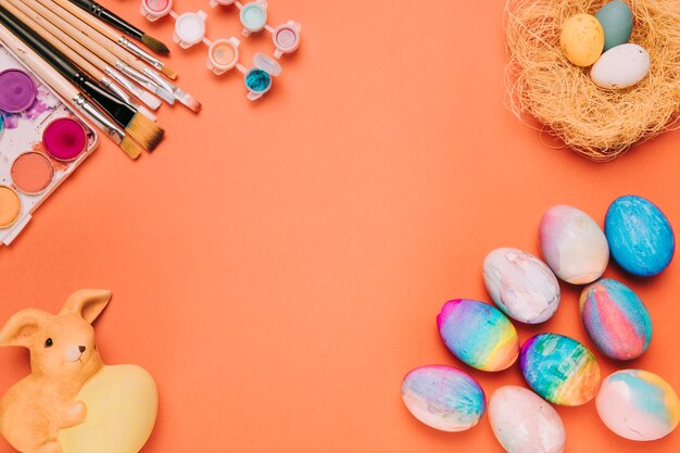 Colorful easter eggs; nest; paint brushes; paint water color box and rabbit statue against orange backdrop