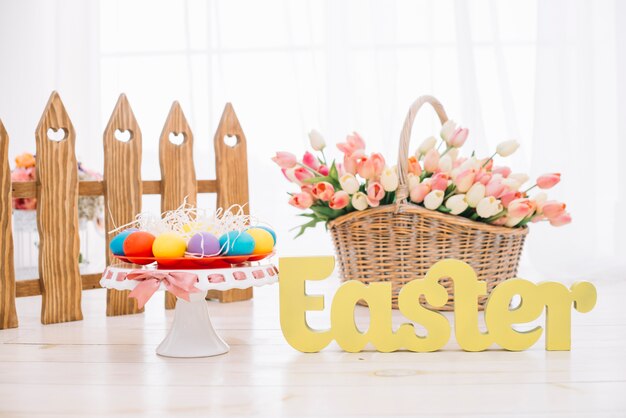 Красочные пасхальные яйца; корзина тюльпанов с желтым пасхальным текстом на деревянном столе