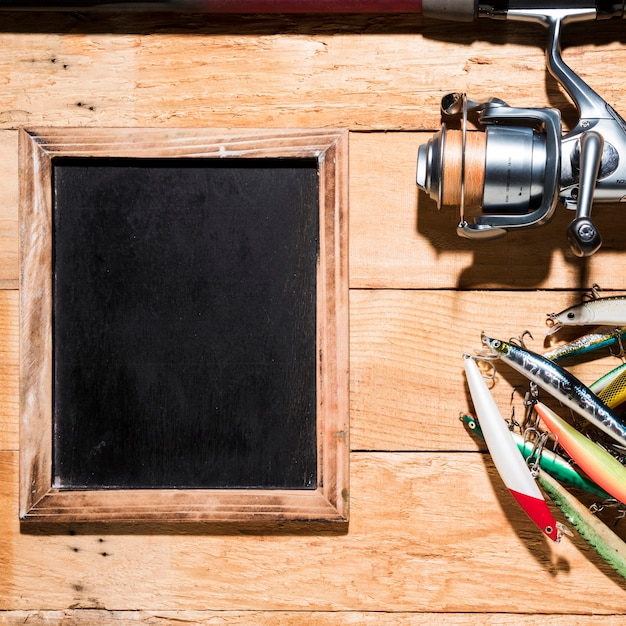 Colorful fishing baits; fishing reel near the blank slate on wooden desk