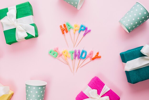 Colorful happy birthday candles surrounded with gift boxes and disposable glasses on pink background