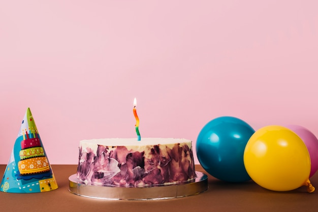 Colorful illuminated candle on birthday cake with party hat and balloons on desk against pink background