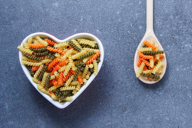 Colorful macaroni pasta in a heart shaped bowl and spoon on a gray surface