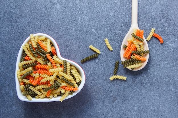 Colorful macaroni pasta in a heart shaped bowl and spoon top view on a gray surface