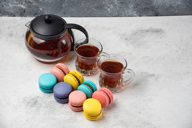 Macarons colorati con tazza da tè e due tazze di tè nero su superficie bianca.