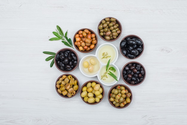 Colorful olives and olive oil with olive leaves in a clay and white bowls on white wood plank, top view.