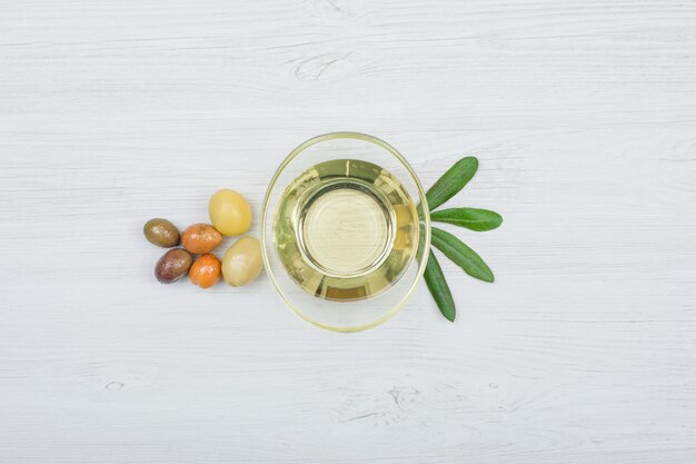 Colorful olives and olive oil with olive leaves in a glass can on white wood plank, top view.