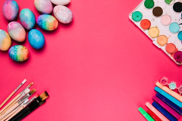 Разноцветные расписные пасхальные яйца; кисти для рисования; коробка для краски и фломастер на розовом фоне