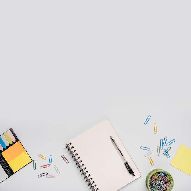 Цветные скрепки; спиральный ноутбук с ручкой и клейкие заметки на белом фоне