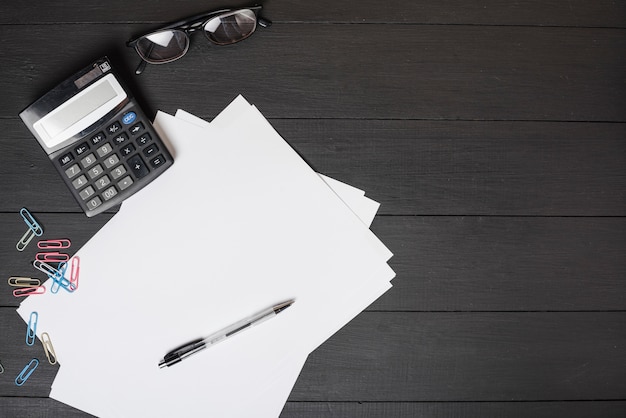 Colorful paper clips; white paper; pen; calculator and sunglasses on black wooden background