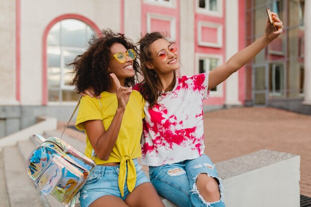 Colorful portrait of happy young girls friends smiling sitting in street taking selfie photo on mobile phone, women having fun together