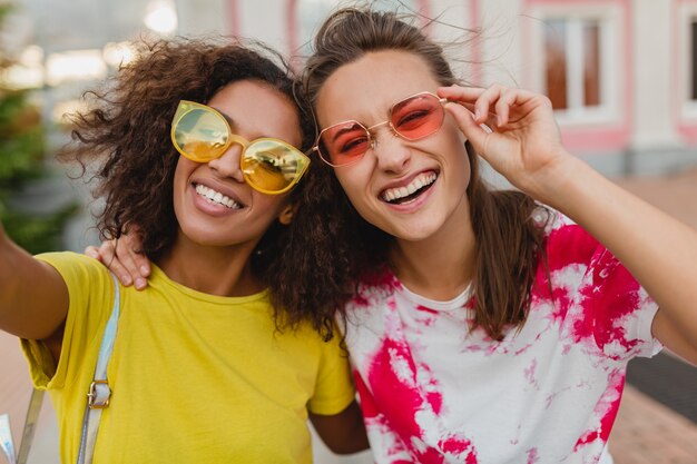 Colorful portrait of happy young girls friends smiling sitting in street taking selfie photo on mobile phone, women having fun together