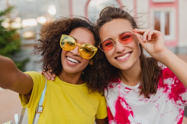 Colorful portrait of happy young girls friends smiling sitting in street taking selfie photo on mobile phone, women having fun together