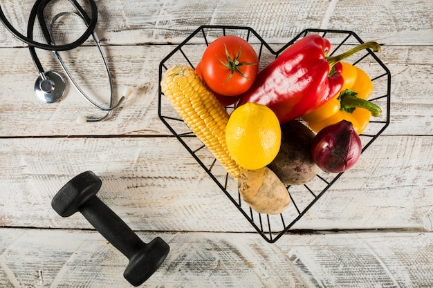 Colorful raw vegetables in container near dumbbell and stethoscope on wooden surface