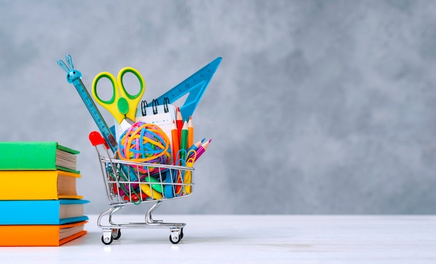 Colorful school supplies in the shopping basket on a gray background with a copy of the text space. A stack of books with colorful covers. The concept of returning to school for the new academic year.