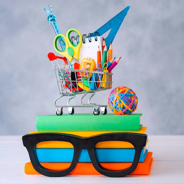 Colorful school supplies shopping basket gray wall with a copy text space. A stack of books with colorful covers frame glasses. The concept of returning to school for new academic year.