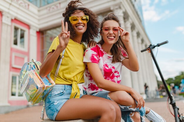 Colorful stylish happy young girls friends smiling sitting in street, women having fun together