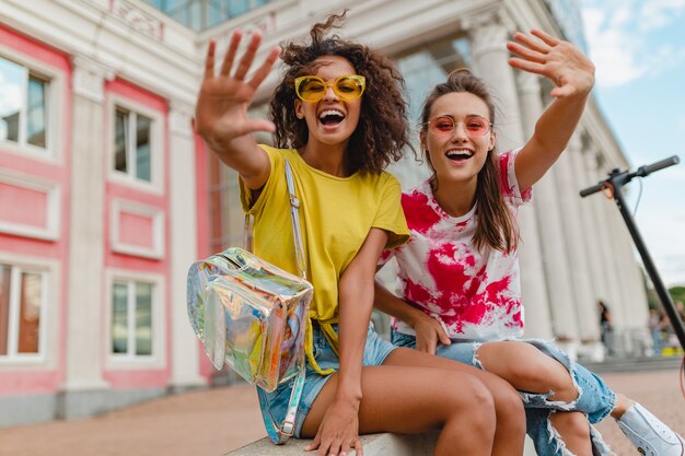 Colorful stylish happy young girls friends smiling sitting in street, women having fun together