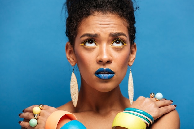 Foto gratuita donna di razza mista entusiasta colorato con trucco alla moda e accessori guardando verso l'alto con le mani incrociate sulle spalle, sul muro blu