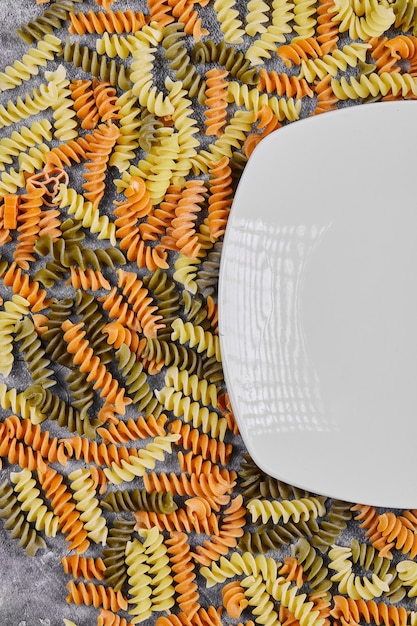 Colorful uncooked fusilli next to white plate on white.