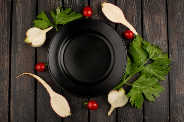 Colorful vegetables fresh ripe red cherry tomatoes and cut radishes with green leafs around black plate on a wooden rustic background