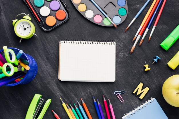 Colourful stationery, paints and clock surrounding notebook on grey background