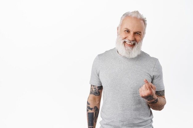 Come closer. Happy handsome old man with tattoos beckon to approach, flick finger, smiling and laughing while luring people, inviting go with him, standing over white background