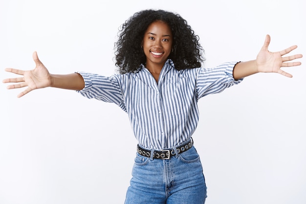 Vieni lasciati abbracciare. ritratto amichevole dall'aspetto attraente amica afroamericana dare un caloroso benvenuto vuole abbracciare coccole, estendere le braccia lateralmente sorridendo ampiamente salutando gli ospiti, muro bianco