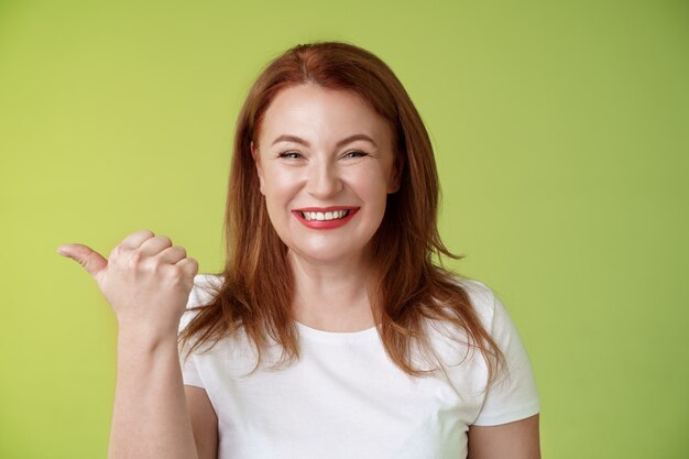 Come visit our store cheerful pleasant friendly charming redhead middleaged woman entrepreneur pointing left thumb stand green wall