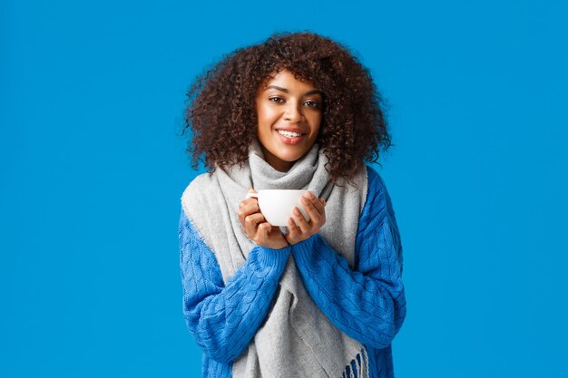 Comfort, tenderness and warmth concept. Charming lovely african american woman with afro haircut, in sweater and scarf, warming up with nice cup tea, enjoying hot coffee.