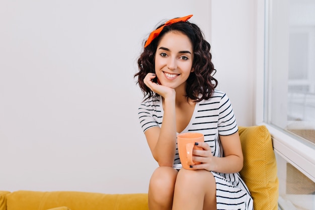 Tempo confortevole a casa con una tazza di tè di giovane donna straordinaria in vestito rilassante sul divano vicino alla finestra in appartamento moderno. felicità, gioia, relax, moda, sorriso