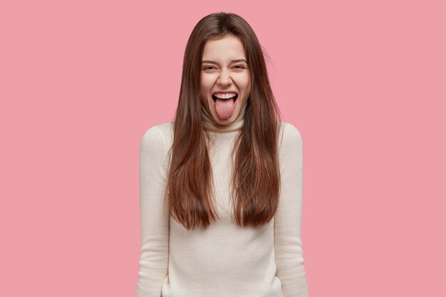 Comic young female model sticks out tongue, makes happy grimace, dressed in white clothes, has long hair, expresses positive emotions