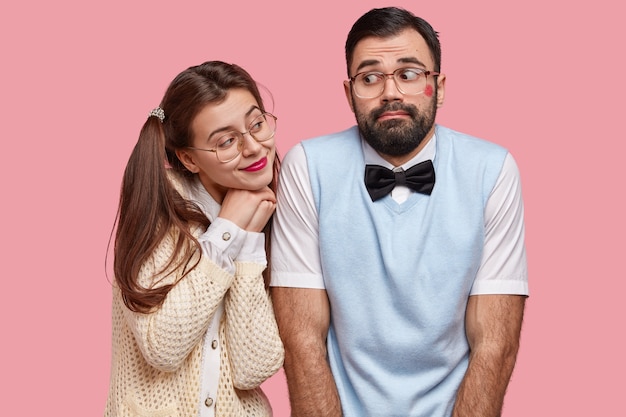 Comic young woman with two pony tails looks with love at boyfriend who has red lipstick on cheek after recieving kiss