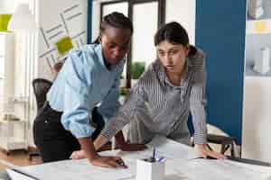 Free photo comitted team of engineers partners discussing construction plans standing at desk in modern architectural office. team of two women architects designing blueprints for real estate housing project.