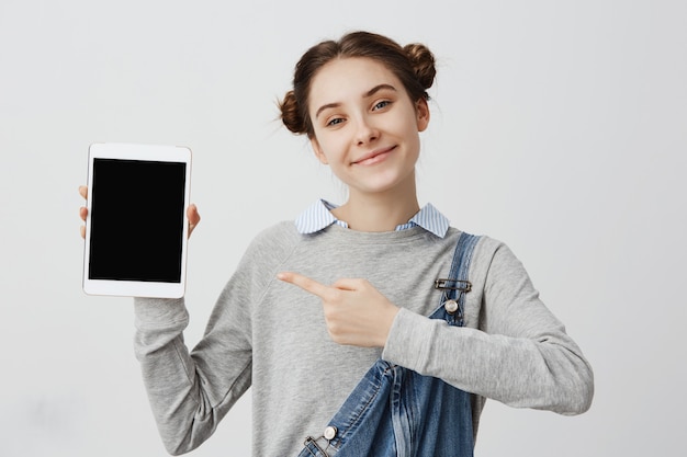 Commercial with beautiful woman smiling while showing switched on screen of tablet gadget. Smart girl with odango buns presenting modern device showing its workability. Marketing, sales