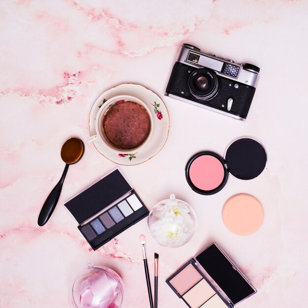 Compact face powder; ribbon; coffee cup; makeup brush; eyeshadow palette and vintage camera on pink textured backdrop