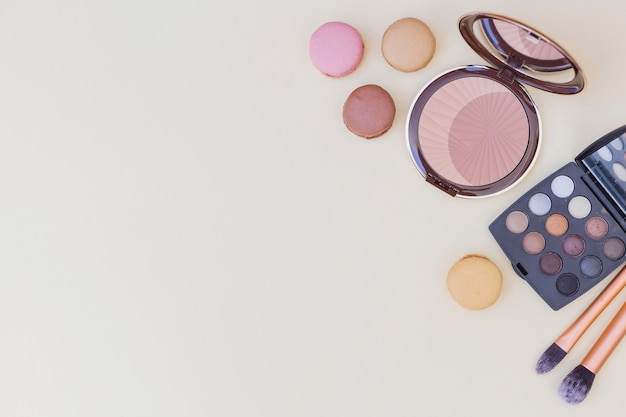 Compact powder; eyeshadow palette; makeup brush and macaroons on beige background