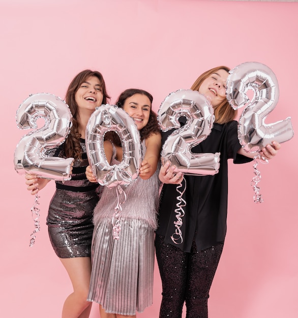 A company of cheerful girlfriends on a pink studio background with silver balloons in the form of the numbers 2022.