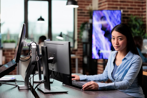 Company office worker in production department working on computer while designing modern graphics. Beautiful asian creative artist sitting at desk while developing digital art and looking at camera.