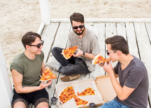 Компания молодых парней ест пиццу на пляже