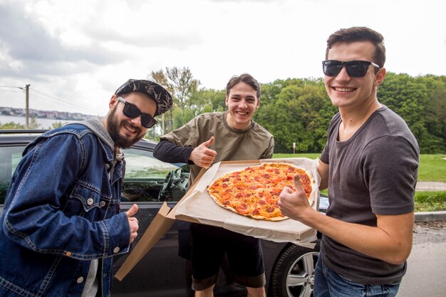 Company of young guys with pizza doing excellent sign in nature