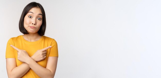 Complicated asian girl pointing fingers sideways showing left and right choices staring indecisive clueless standing over white background