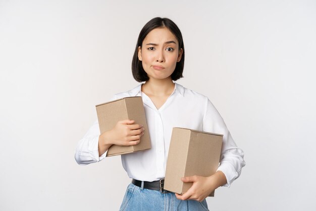 Complicated young asian woman holding two boxes looking doubtful at camera standing over white background puzzled