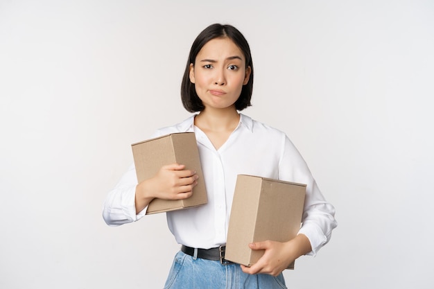 Сложная молодая азиатская женщина, держащая две коробки, с сомнением смотрит в камеру, стоящую на белом фоне, озадаченная
