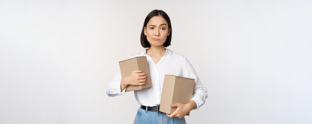 Complicated young asian woman holding two boxes looking doubtful at camera standing over white background puzzled