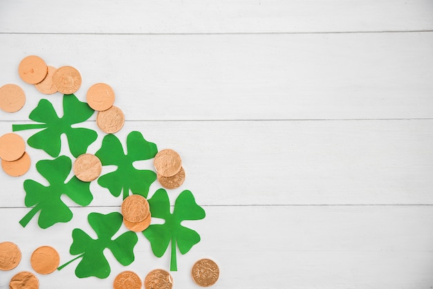 Composition of heap of coins and green paper clovers on board