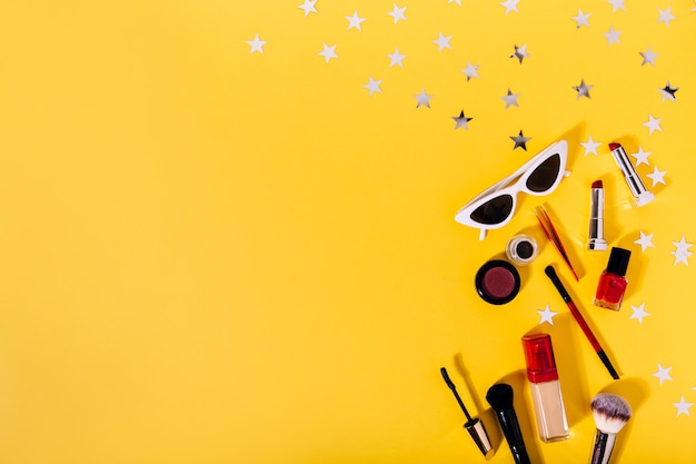 Composition of makeup brushes, tonal foundation, eye liner, lipstick, mascara and stylish sunglasses on orange wall with silver stars