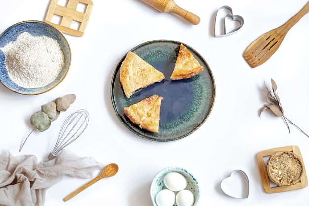 Composition with a pieces of pie on a plate and ingredients for cooking and kitchen accessories on white table.